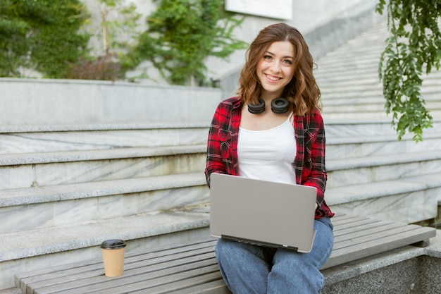 Young cheerful woman student using laptop outdoors. Distance work or education concept