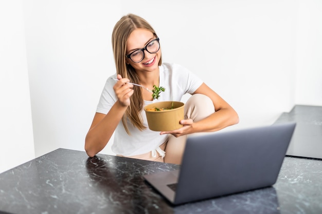 Young cheerful woman using on laptop computer and eating salad in the kitchen