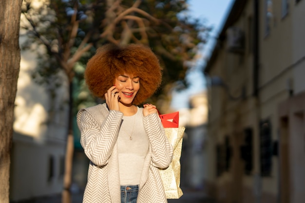 Young cheerful woman with afro hair holding shopping bags and talking on smartphone in the city street