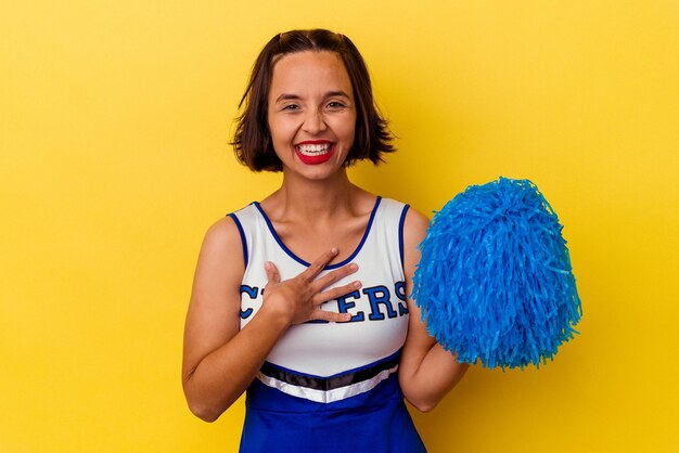 Young cheerleader mixed race woman isolated on yellow background laughs out loudly keeping hand on chest.