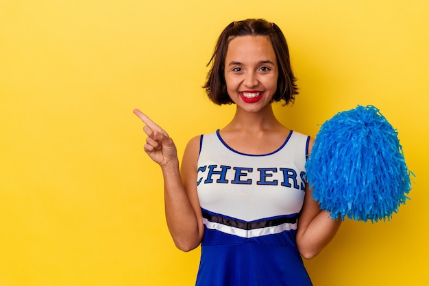 Young cheerleader mixed race woman isolated on yellow wall smiling and pointing aside, showing something at blank space