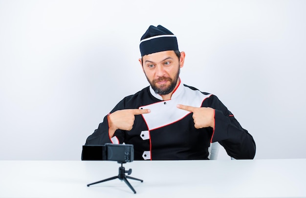 Young chef blogger is posing to his mini camera by pointing himself with forefingers on white background