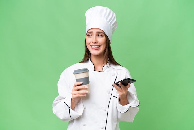 Young chef caucasian woman over isolated background holding coffee to take away and a mobile while thinking something