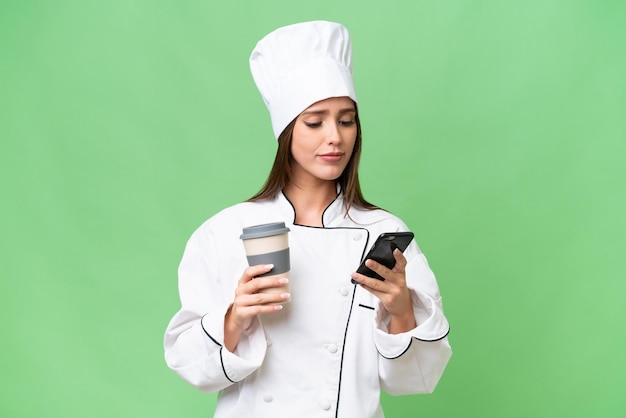 Young chef caucasian woman over isolated background holding coffee to take away and a mobile