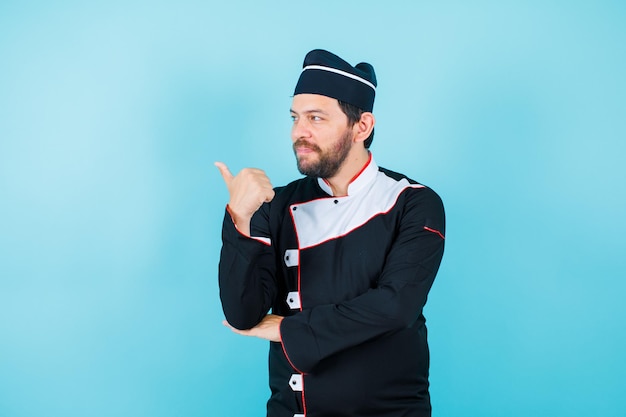 Young chef is looking left side and showing back with thumb on blue background