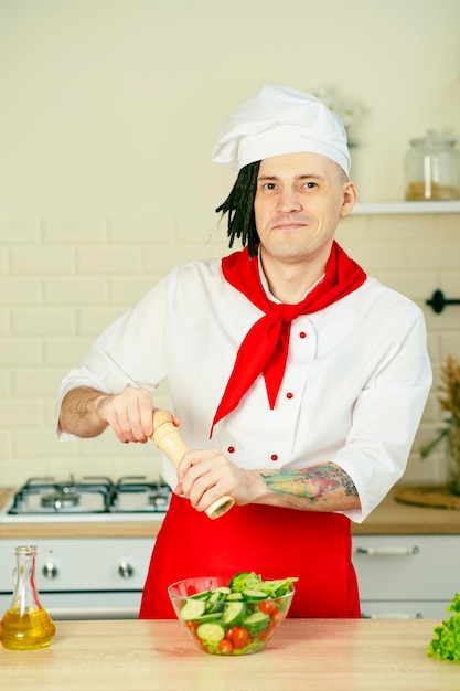 Young chef with dreadlocks peppering vegetable salad in kitchen Smiling man in uniform preparing salad grinding spices with mill