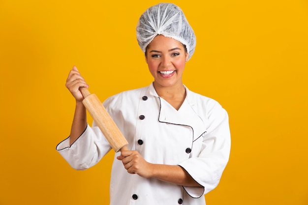 Young chef woman in white uniform holds rolling pin on yellow background