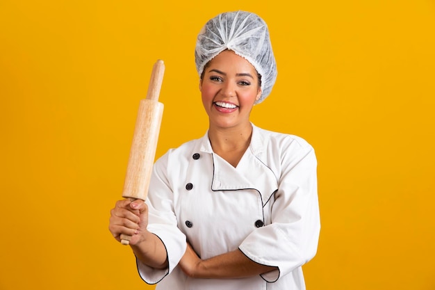 Young chef woman in white uniform holds rolling pin on yellow background