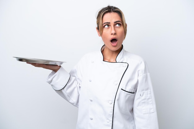 Young chef woman with tray isolated on white background looking up and with surprised expression