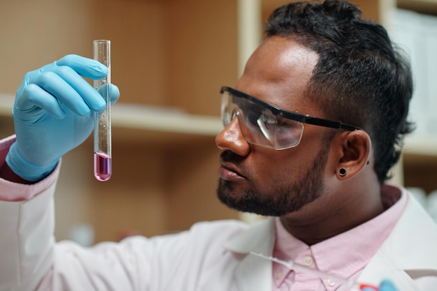 Young chemist in gloves eyeglasses and lab coat looking at flask containing liquid substance while s