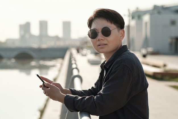 Young Chinese businesswoman in sunglasses standing by riverside and scrolling in smartphone