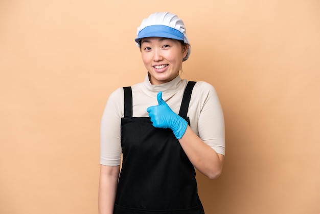 Young Chinese Fishmonger woman wearing an apron and holding a raw fish isolated on pink background giving a thumbs up gesture