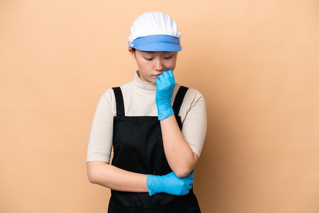 Young Chinese Fishmonger woman wearing an apron and holding a raw fish isolated on pink background having doubts