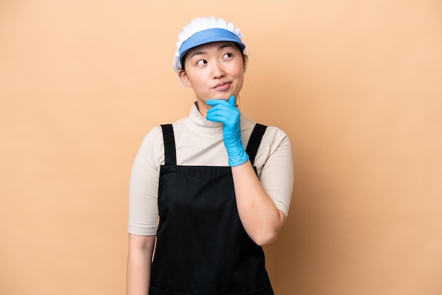 Young Chinese Fishmonger woman wearing an apron and holding a raw fish isolated on pink background and looking up