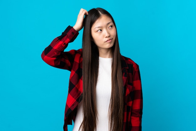 Young Chinese girl over isolated blue wall having doubts while scratching head