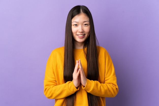 Young Chinese girl over isolated purple wall keeps palm together. Person asks for something