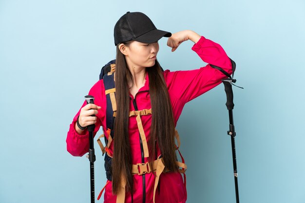 Young Chinese girl with backpack and trekking poles over blue doing strong gesture