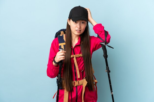 Young Chinese girl with backpack and trekking poles over blue wall doing nervous gesture