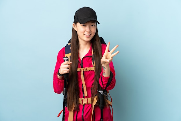 Photo young chinese girl with backpack and trekking poles over isolated blue wall happy and counting three with fingers