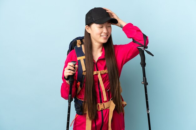 Young Chinese girl with backpack and trekking poles over isolated blue wall smiling a lot