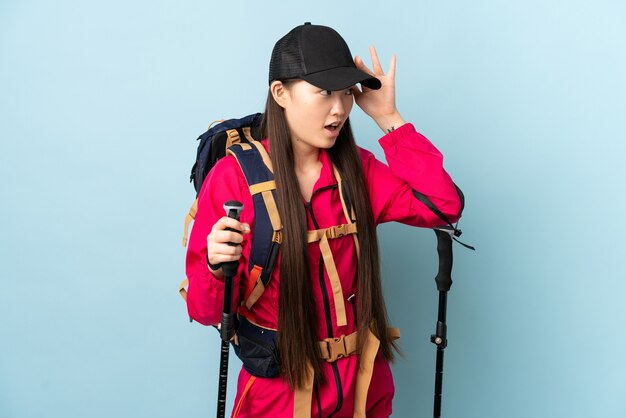 Young Chinese girl with backpack and trekking poles on isolated blue with surprise expression while looking side