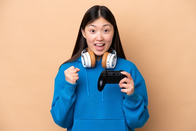 Young Chinese man playing with a video game controller isolated on beige background celebrating a victory in winner position