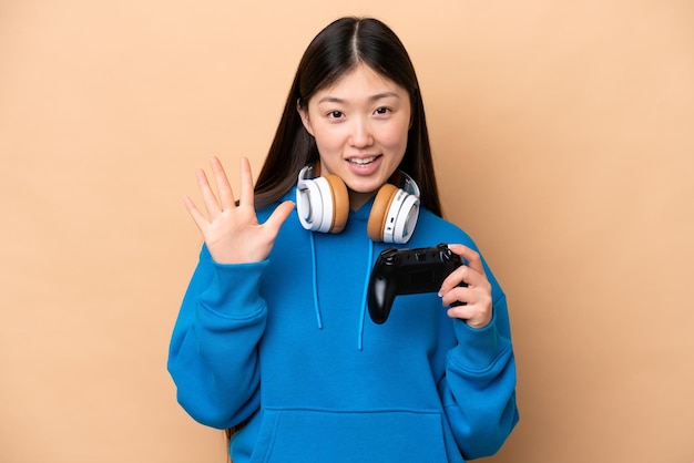 Young Chinese man playing with a video game controller isolated on beige background counting five with fingers