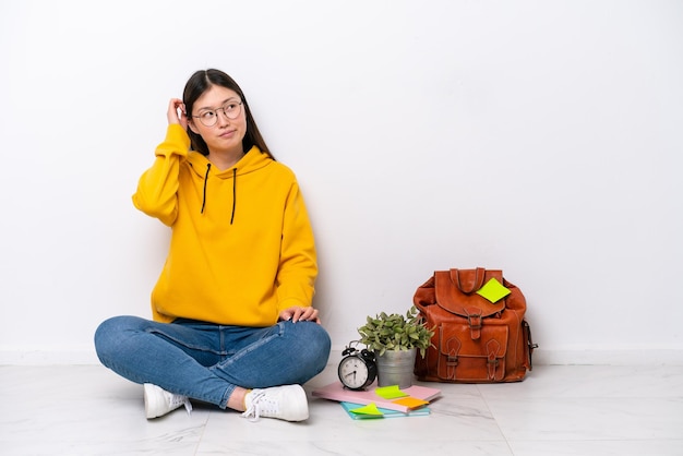 Young Chinese student woman sitting on the floor isolated on white wall having doubts and with confuse face expression