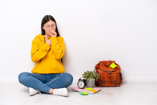 Young Chinese student woman sitting on the floor isolated on white wall is suffering with cough and feeling bad