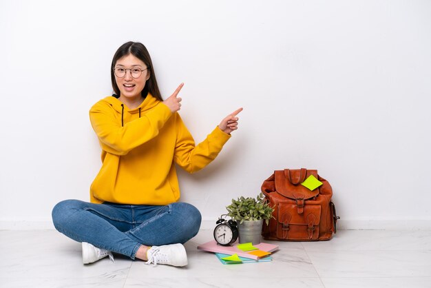 Young Chinese student woman sitting on the floor isolated on white wall pointing finger to the side and presenting a product