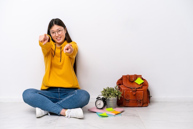 Young Chinese student woman sitting on the floor isolated on white wall points finger at you while smiling