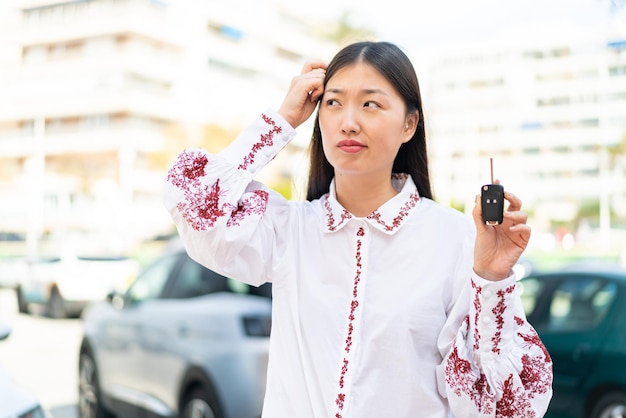 Young Chinese woman holding car keys at outdoors having doubts and with confuse face expression