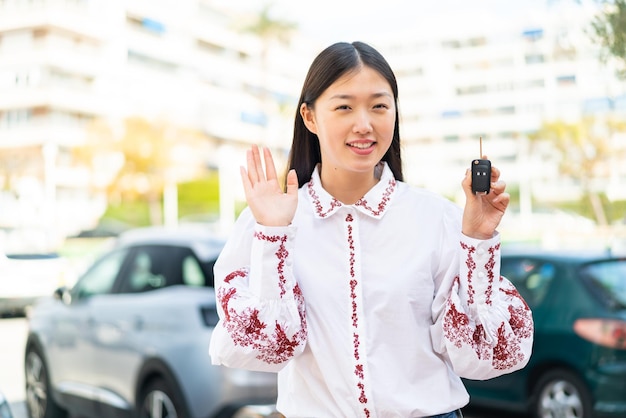 Young Chinese woman holding car keys at outdoors saluting with hand with happy expression