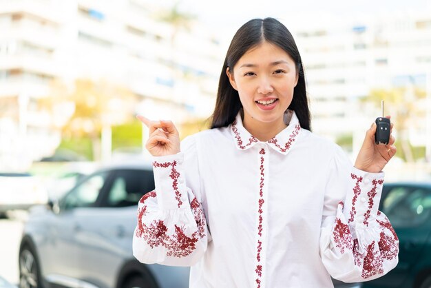 Young Chinese woman holding car keys at outdoors surprised and pointing finger to the side