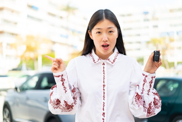 Young Chinese woman holding car keys at outdoors surprised and pointing side