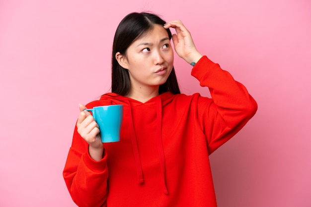 Young Chinese woman holding cup of coffee isolated on pink background having doubts and with confuse face expression