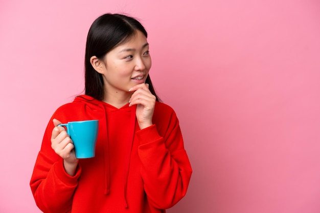 Young Chinese woman holding cup of coffee isolated on pink background thinking an idea and looking side