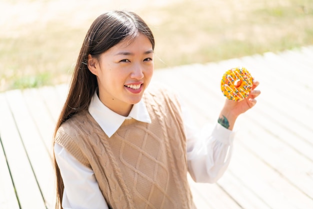 Young Chinese woman holding a donut at outdoors smiling a lot