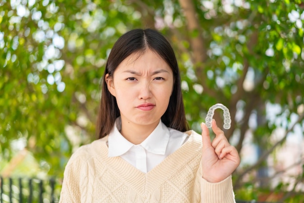 Young Chinese woman holding invisible braces at outdoors with sad expression