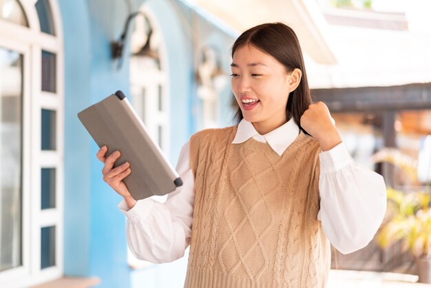 Young Chinese woman holding a tablet at outdoors celebrating a victory