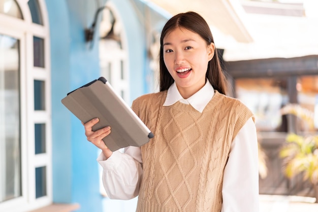 Young Chinese woman holding a tablet at outdoors with surprise and shocked facial expression