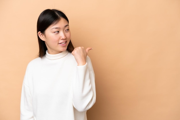 Young Chinese woman isolated on beige background pointing to the side to present a product