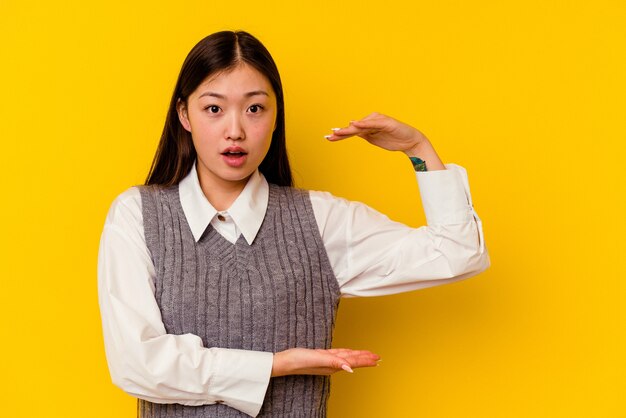 Young chinese woman isolated on yellow shocked and amazed holding a copy space between hands.