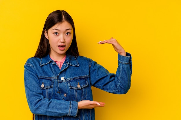 Young chinese woman isolated on yellow shocked and amazed holding a copy space between hands.