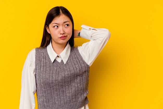 Young chinese woman isolated on yellow touching back of head, thinking and making a choice.