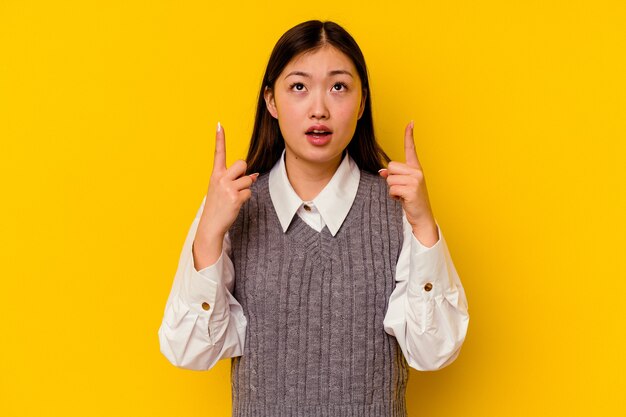 Young chinese woman isolated on yellow wall pointing upside with opened mouth.