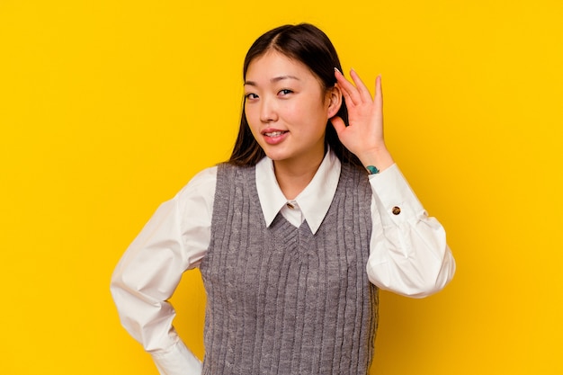 Young chinese woman isolated on yellow wall trying to listening a gossip.