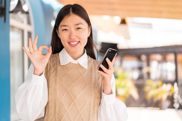 Young Chinese woman at outdoors using mobile phone and doing OK sign