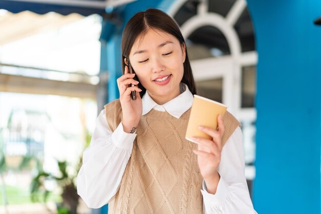 Young Chinese woman at outdoors using mobile phone and holding a coffee with happy expression