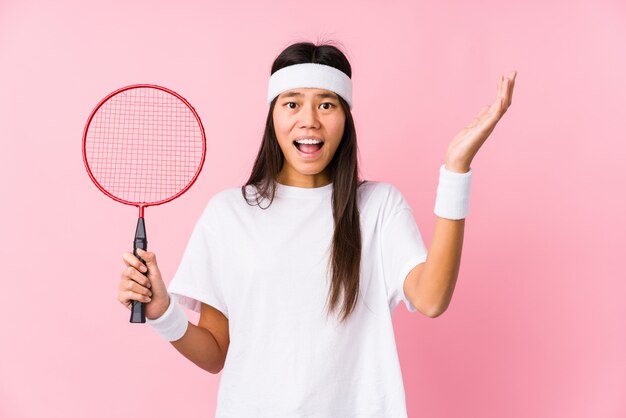 Young chinese woman playing badminton in a pink background receiving a pleasant surprise, excited and raising hands.